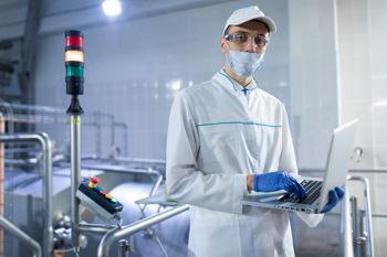 Person in clean white lab coat and blue latex gloves looking at a laptop in a food processing area