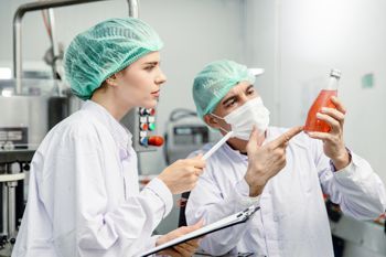 Two workers in clean white gear examining a colored liquid in a glass bottle