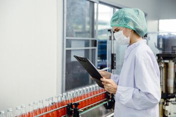 A worker in clean white gear examining a conveyor line of clear glass bottles